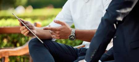 Two businessmen on a bench outside discussing business whilst overlooking a tablet.