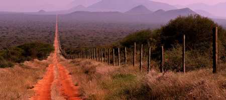 A red dirt road in the middle of the countryside, with a wooden fence running alongside it.